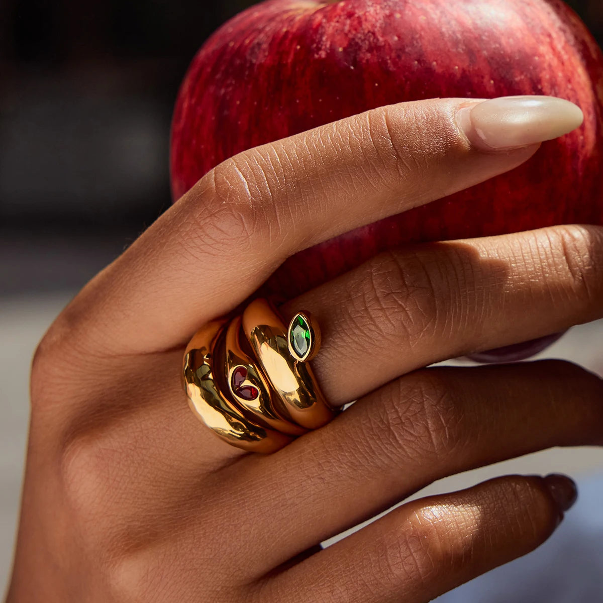 Hand wearing a gold ring with gemstones holding a red apple.