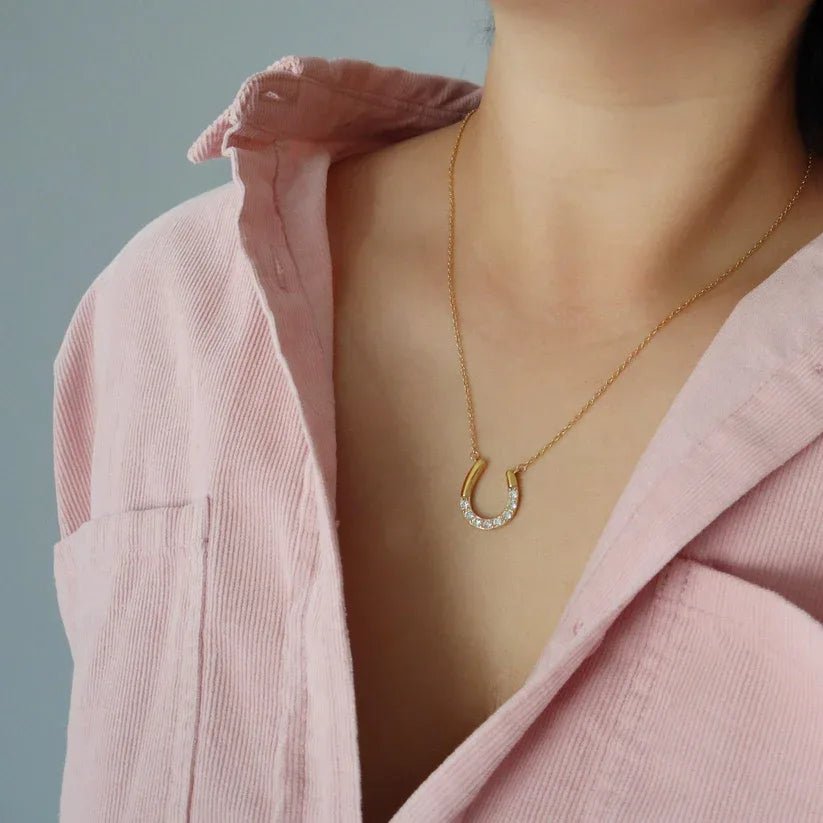 Person wearing a gold necklace with a horseshoe pendant against a neutral background